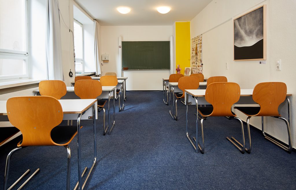 Unterrichtsraum mit Tischreihen. An der hinteren Wand eine Tafel und anatomische Grafiken. Blauer Teppichboden