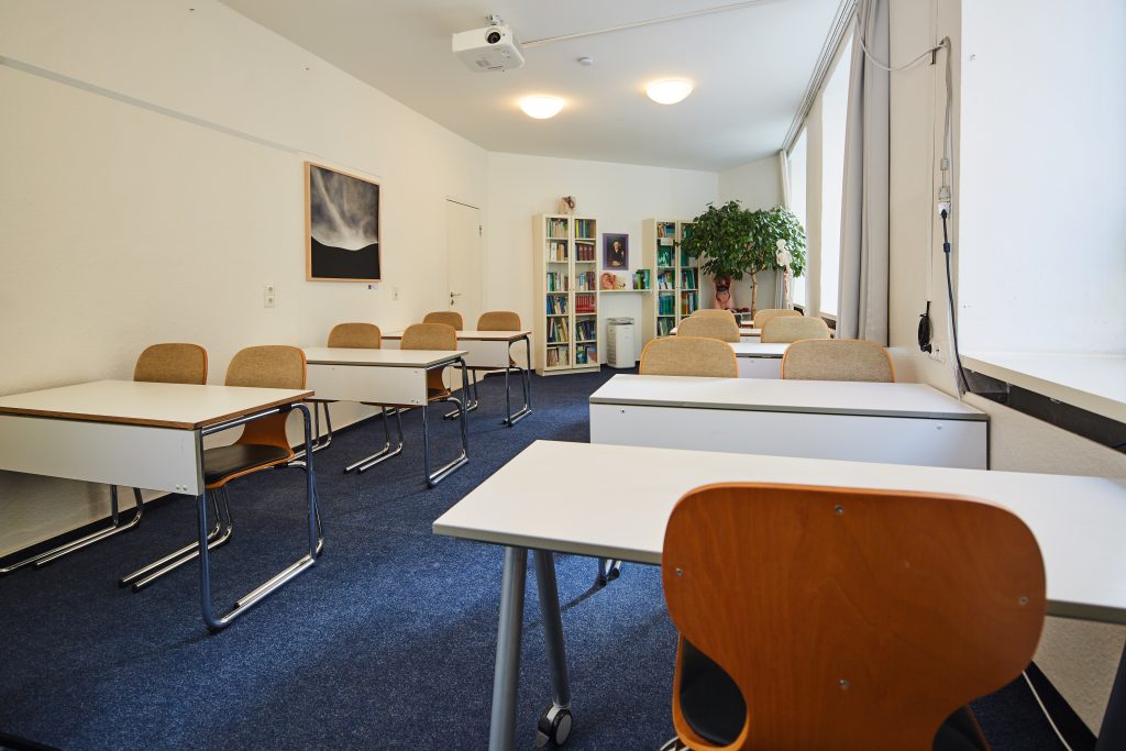 Unterrichtsraum mit Tischreihen. An der hinteren Wand weiße Bücherregale und eine Pflanze. Blauer Teppichboden